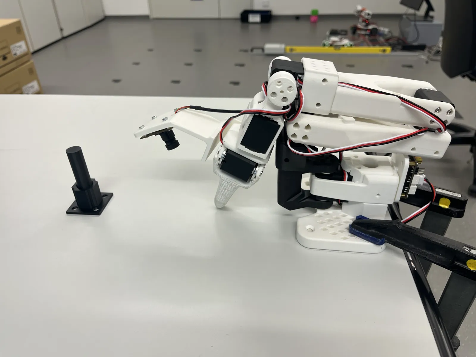 SO-101 follower arm with wrist camera above a white table, with the black peg fixture in front of it